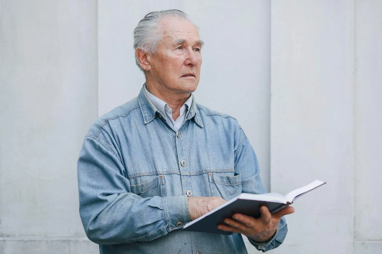 An older man in a denim shirt standing by a pale wall, holding an open book